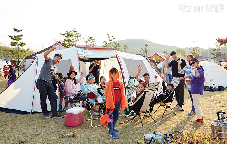 캠핑에 참여한 가족이 텐트 앞에 모여 앉아 카메라를 향해 손을 흔드는 모습.